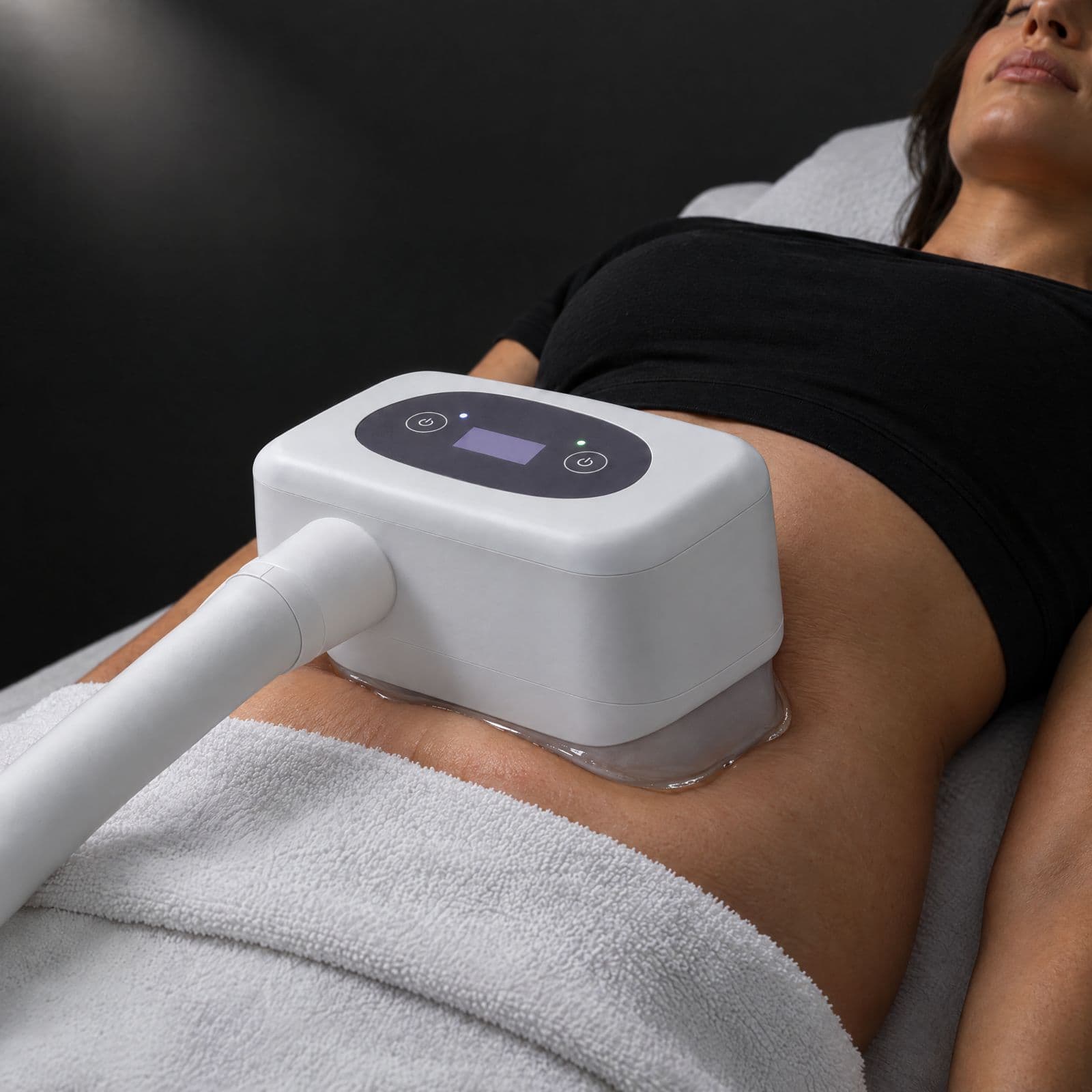 A white cooling applicator rests on a reclining client's gel-covered abdomen, a towel folded over her hips