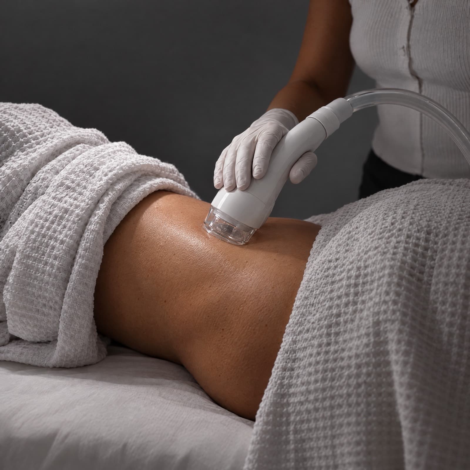 Gloved esthetician passes a clear-tipped vacuum handpiece over a towel-draped client's hip on a treatment bed