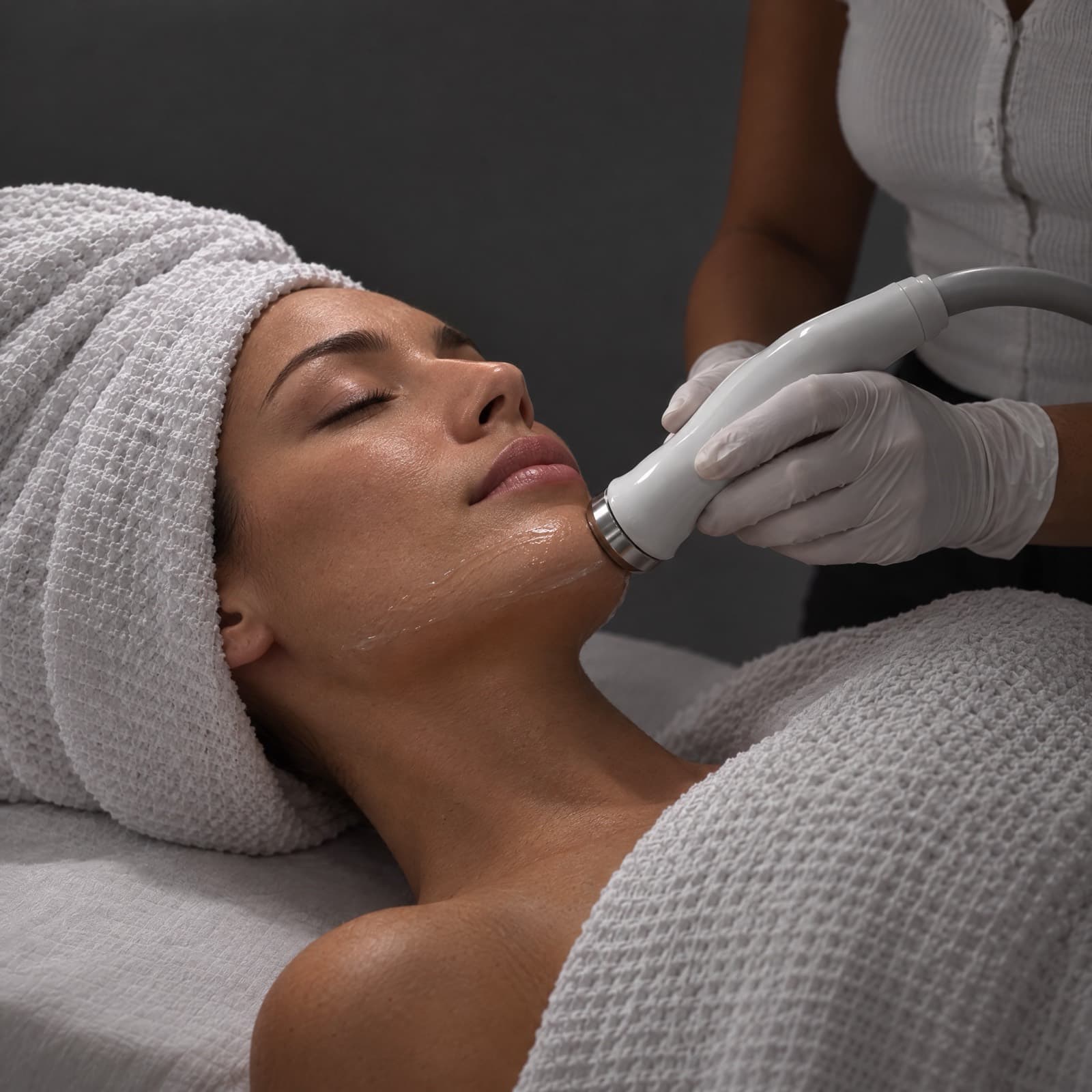 Gloved esthetician guides a slim handpiece along a towel-wrapped client's gel-covered jawline on a treatment bed